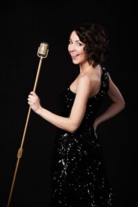 Beautiful young female vocalist in shiny black evening dress holding golden vintage microphone, playful smile, preparing to sing during musical show